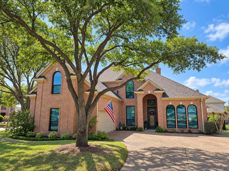 A brick home beneath mature trees with dark-framed arched and rectangular windows.